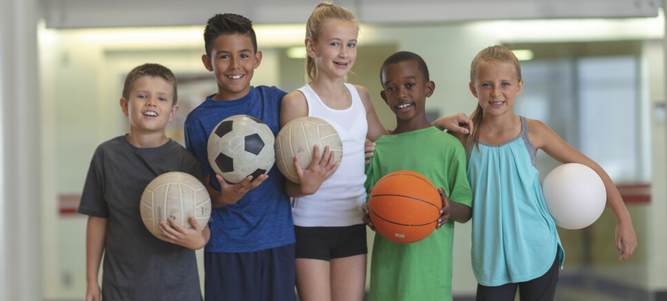 Five kids are standing side-by-side in a gym. They are smiling at the camera and holding sports balls. There is a basketball, soccer ball, two volleyballs, and a dodgeball.